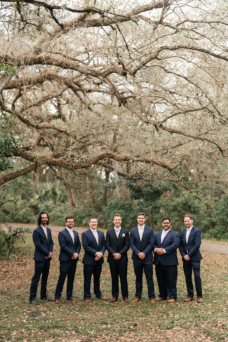 Seven groomsmen in navy suits lined up outdoors beneath a sprawling live oak, smiling at the camera.