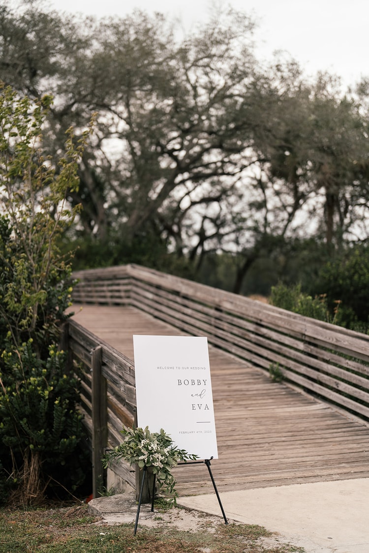 Wedding welcome sign reading "Bobby and Eva, February 4th, 2023" on a white easel at the entrance of the outdoor venue boardwalk.