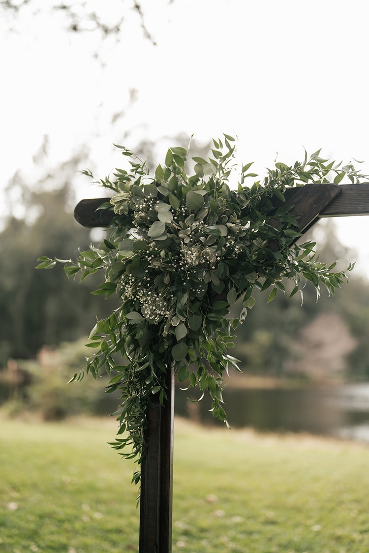 Lush greenery and baby's breath cascade from a dark wood ceremony arch, with a waterway visible in the soft-focus background.