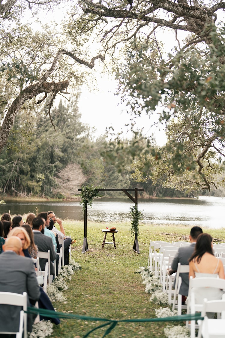 Outdoor ceremony wide view: guests seated in white chairs face a dark wood arch adorned with greenery, set against a still lake.