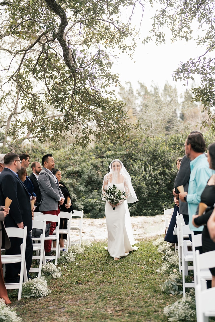 Bride walks alone down the outdoor ceremony aisle beneath a lace-edged veil, guests smiling as she approaches.