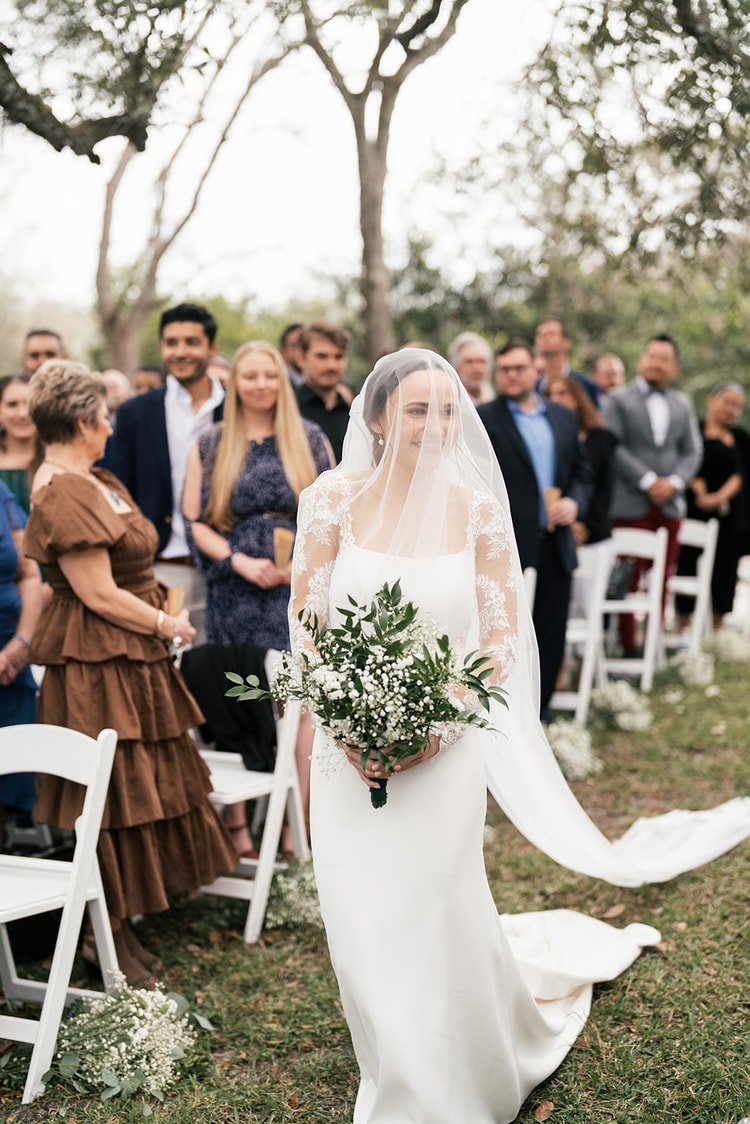Bride walks down the outdoor aisle beneath her cathedral veil, bouquet in hand, with guests standing on both sides.