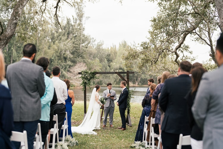 Ceremony wide shot: couple and officiant at the altar beneath the wood arch, guests standing on both sides as vows are exchanged.