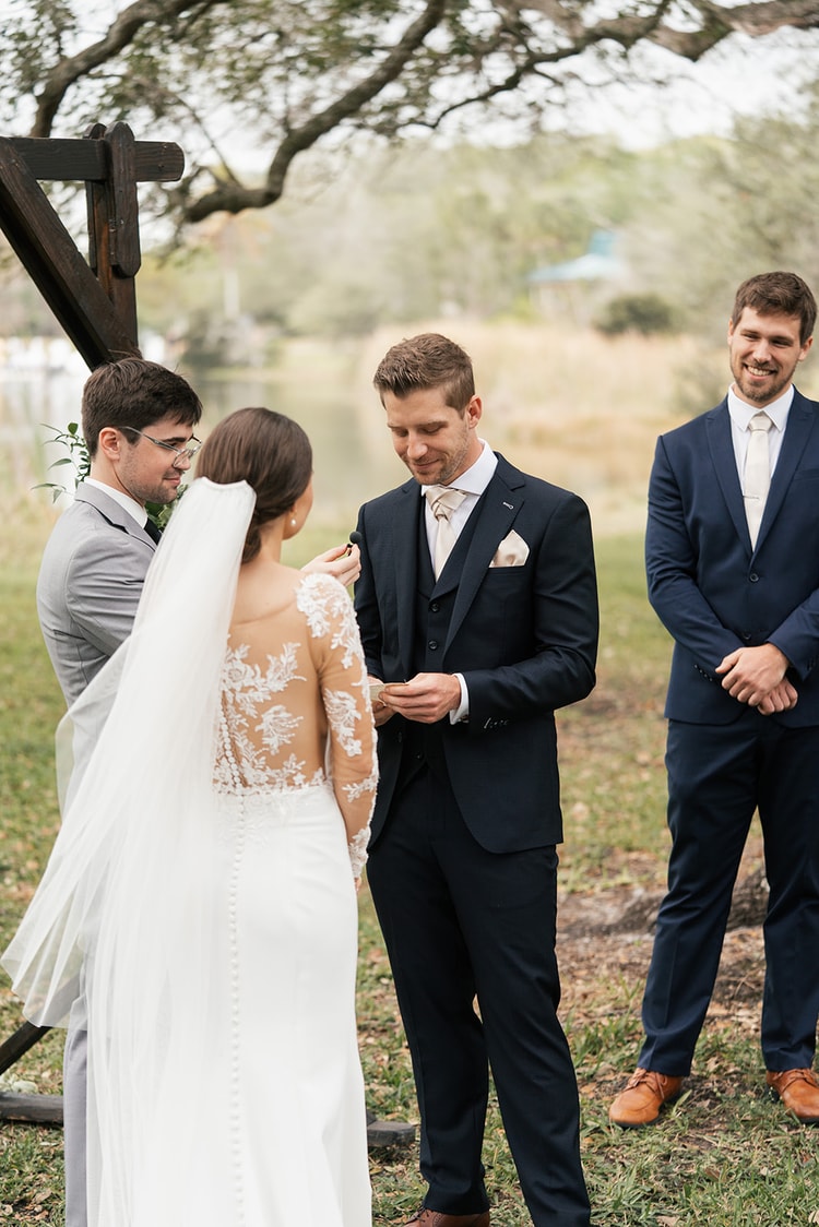 Groom reads his vows at the ceremony altar while the bride faces him, officiant and best man alongside, waterway in the background.