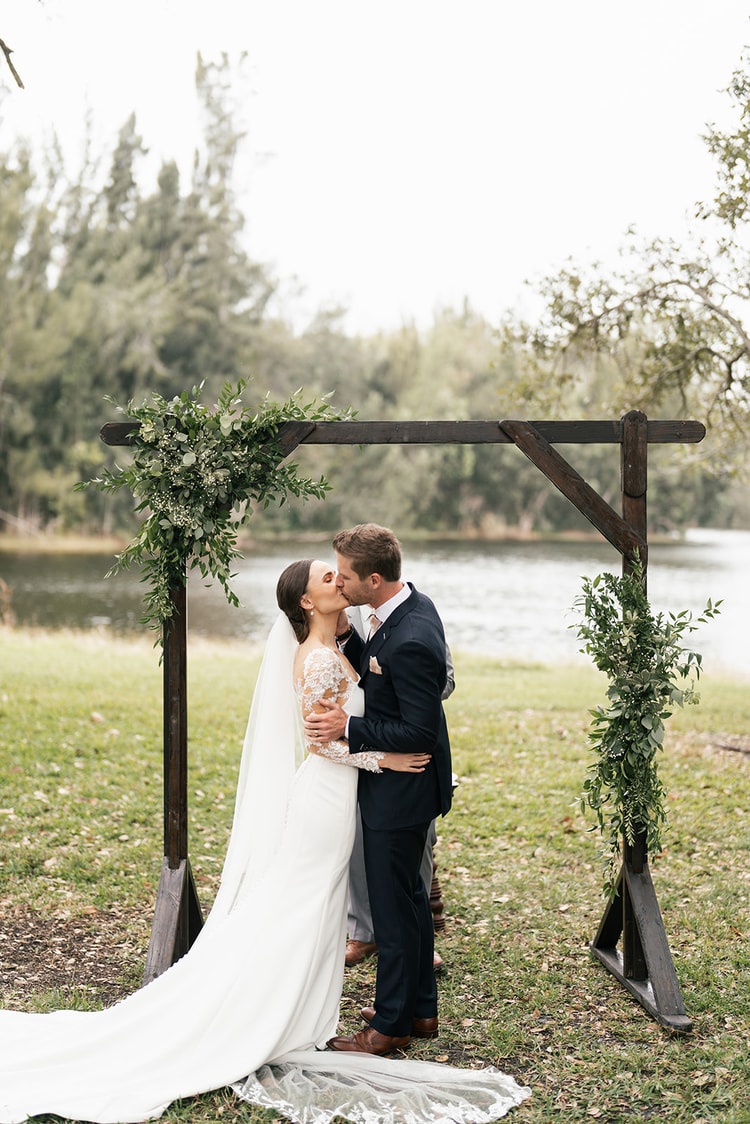 Newlyweds share their first kiss beneath a greenery-draped wooden arch beside a lake, veil trailing behind the bride.