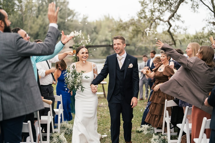 Newlyweds walk back up the aisle as guests toss flower petals, both beaming with wide smiles during the recessional.