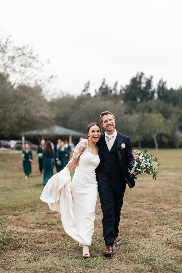Couple walks through a grassy park field laughing together after the ceremony, bridesmaids in teal visible in the background.