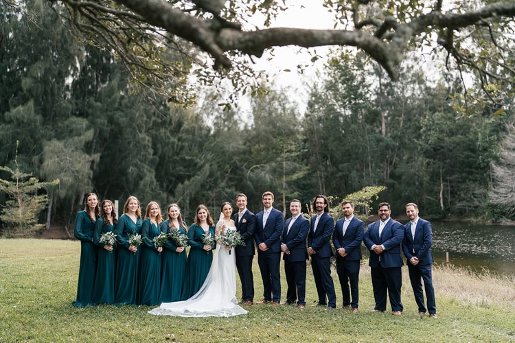 Full wedding party portrait: six bridesmaids in teal gowns, bride, groom, and groomsmen in navy lined up on a lawn beside a pond.