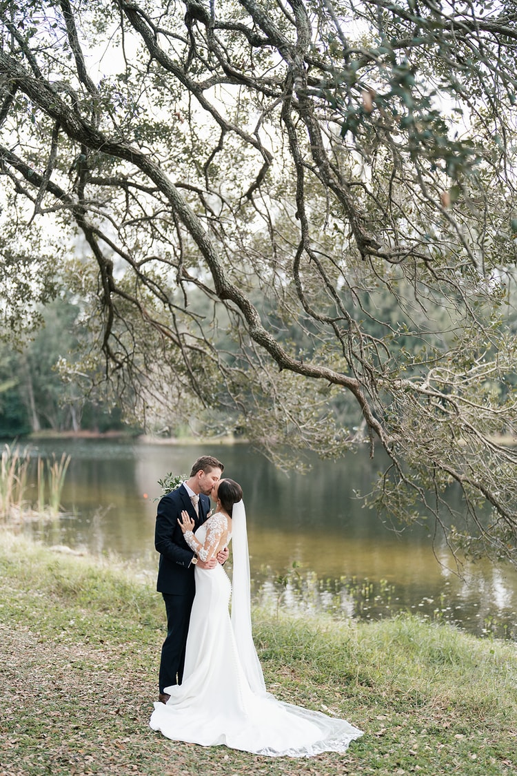 Bride and groom share a quiet kiss beneath a large oak tree by a still pond, veil and train spread around them.