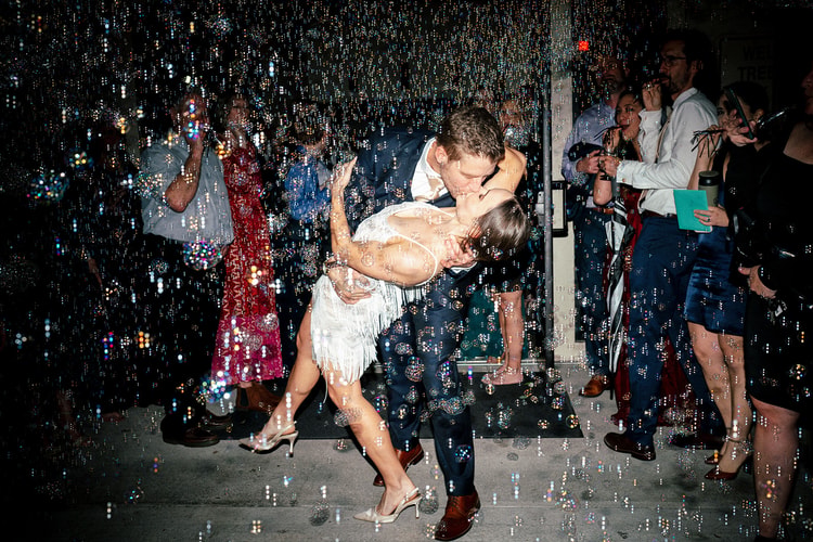 Bride and groom share a dramatic dip kiss on the dance floor as bubbles fill the air and guests celebrate around them.