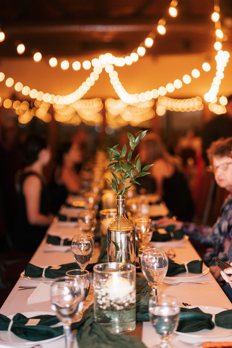 Long reception banquet table set with green linen napkins and greenery stem centerpieces glowing under warm string lights.