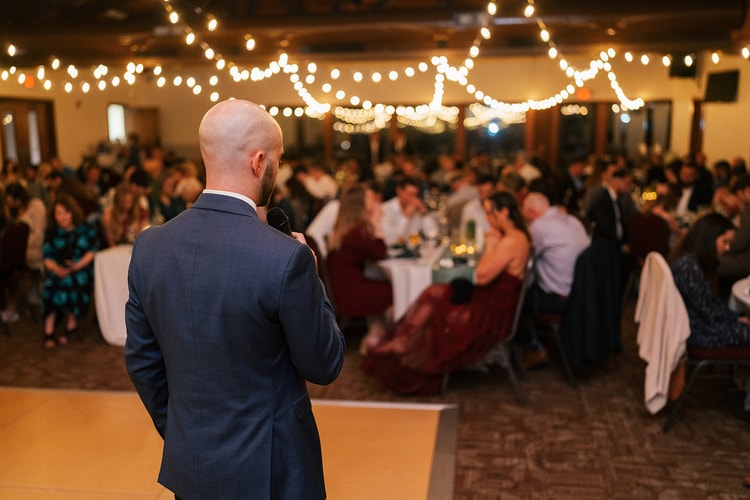 A guest delivers a toast from the dance floor to a full reception room of seated guests glowing under warm bistro lights.