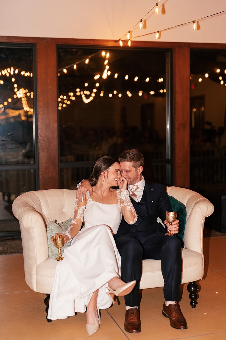 Bride and groom seated together on a cream loveseat under string lights, laughing quietly with gold chalices in hand.