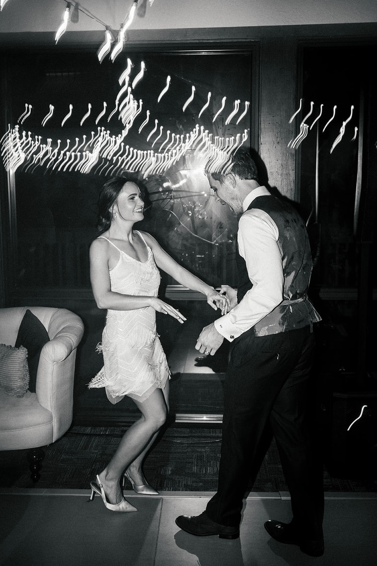 Bride in a fringed mini dress and groom dance and spin together near a large window with reflected string lights, black and white.