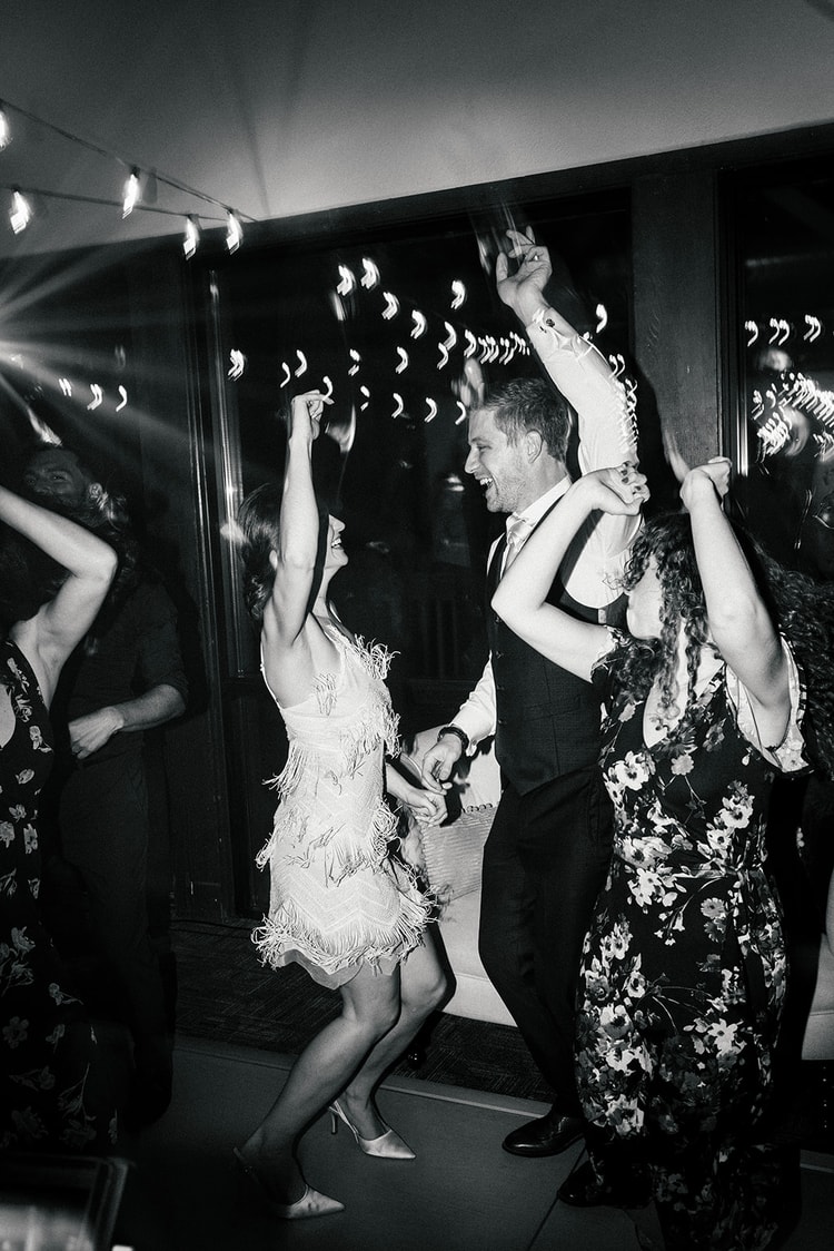 Bride and groom dance energetically with arms raised under reception lighting, guests around them, black and white.