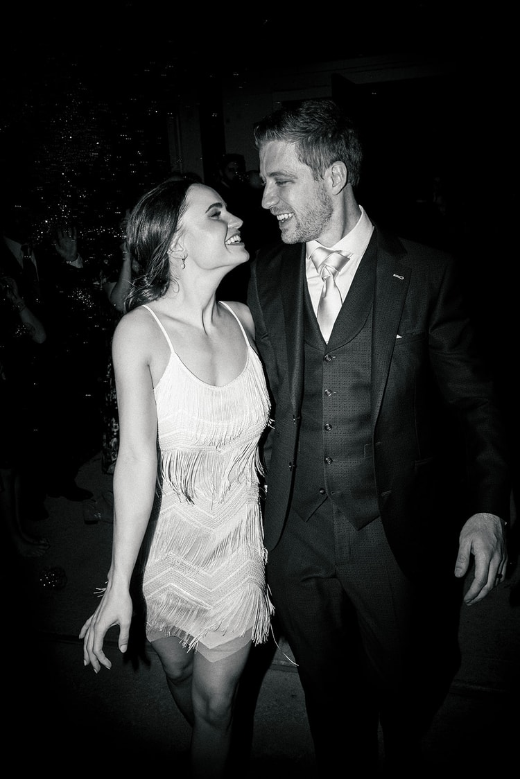 Bride in her fringed reception dress and groom in navy suit laugh at each other on the dance floor, black and white.