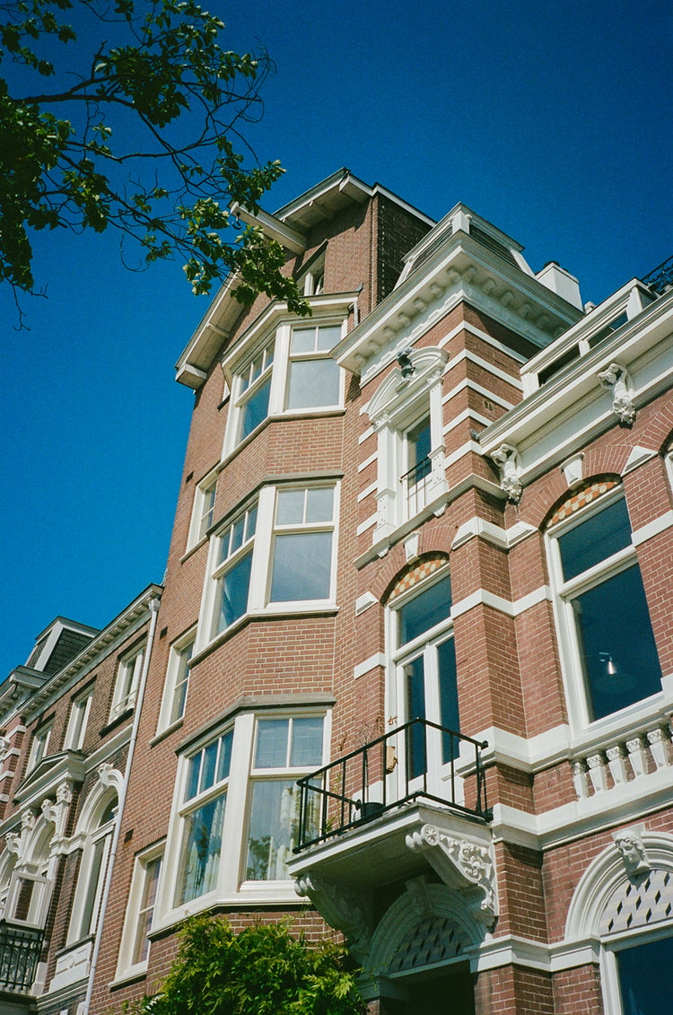 Ornate brick Amsterdam townhouse facade with white-trimmed bay windows and a wrought-iron balcony against a clear blue sky.