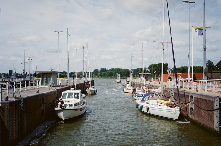 Small boats pass through a canal lock in the Netherlands, sailboats moored along the concrete walls under a cloudy sky.