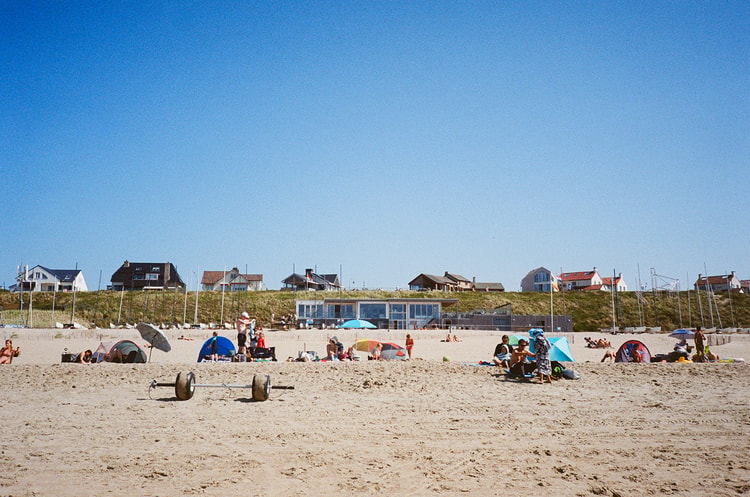 Sunbathers and beach tents dot the sand at a Dutch coastal beach club beneath dune houses and a clear sky.