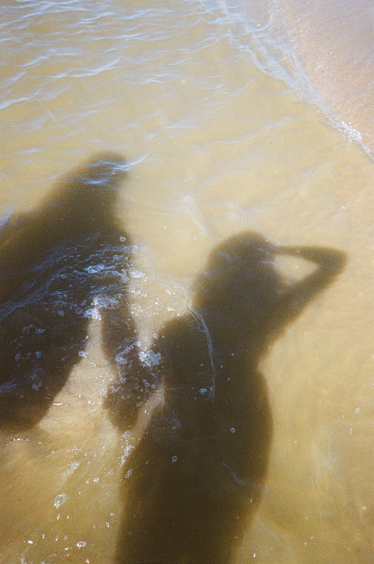 Two blurred shadow silhouettes of a couple stand in shallow, sunlit tidal water on a Dutch beach.