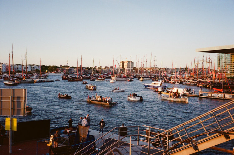 Crowds of small boats gather on Amsterdam's IJ waterway at sunset during a summer sailing festival, tall ship masts in the distance.