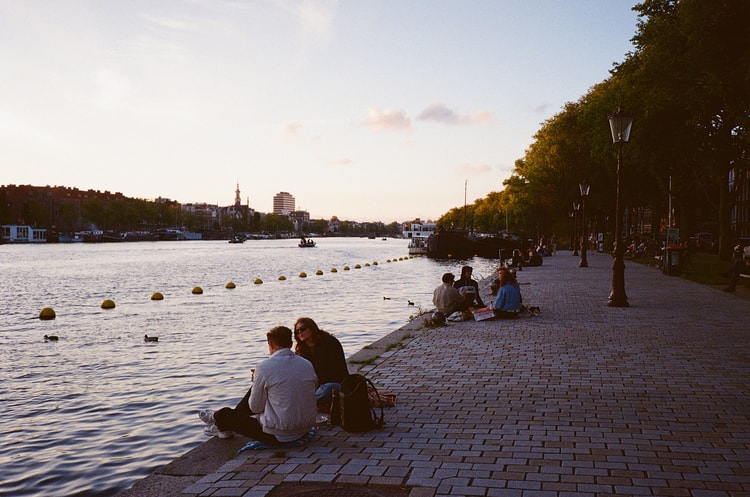 People relax along the edge of an Amsterdam canal at sunset, ducks swimming nearby and a passenger boat gliding past.