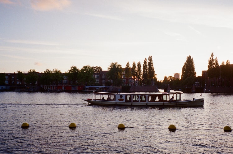 A canal tour boat glides across the Amstel river at sunset, rowhouses and trees silhouetted along the far bank.