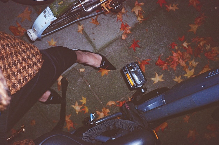Overhead view of a bicycle pedal and a hand adjusting a shoe strap on a leaf-covered Amsterdam pavement in autumn.