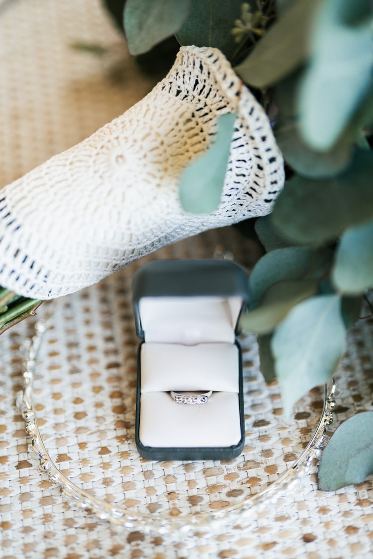 Diamond engagement ring in an open velvet box, surrounded by eucalyptus greenery and a lace bouquet wrap on a rattan surface.