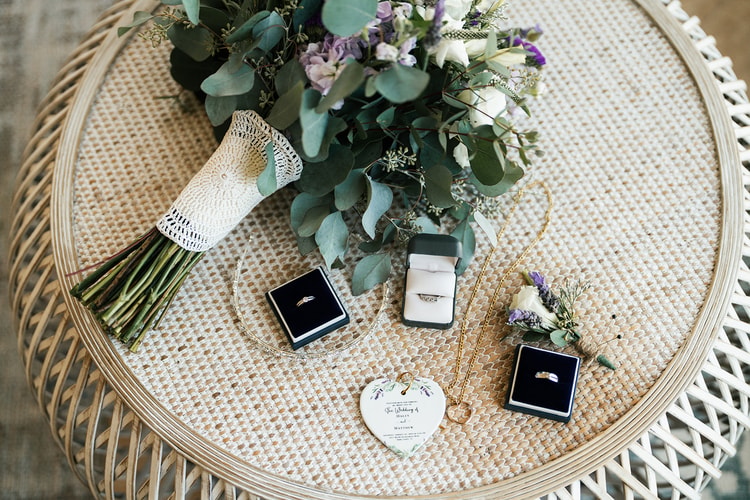 Wedding flat lay showing the bridal bouquet, two ring boxes, boutonniere, heart ornament, and a gold necklace arranged on a rattan side table.