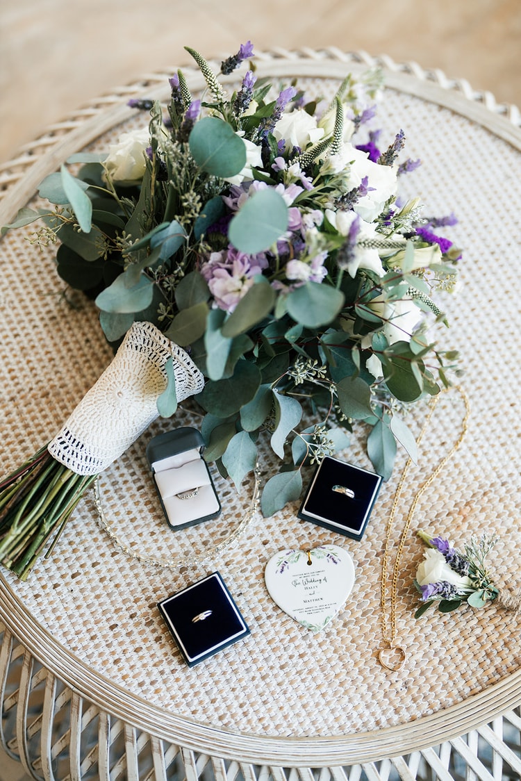 Close-up detail flat lay of a purple and white bridal bouquet with lavender, lisianthus, and eucalyptus alongside two velvet ring boxes and a delicate necklace on a woven surface.