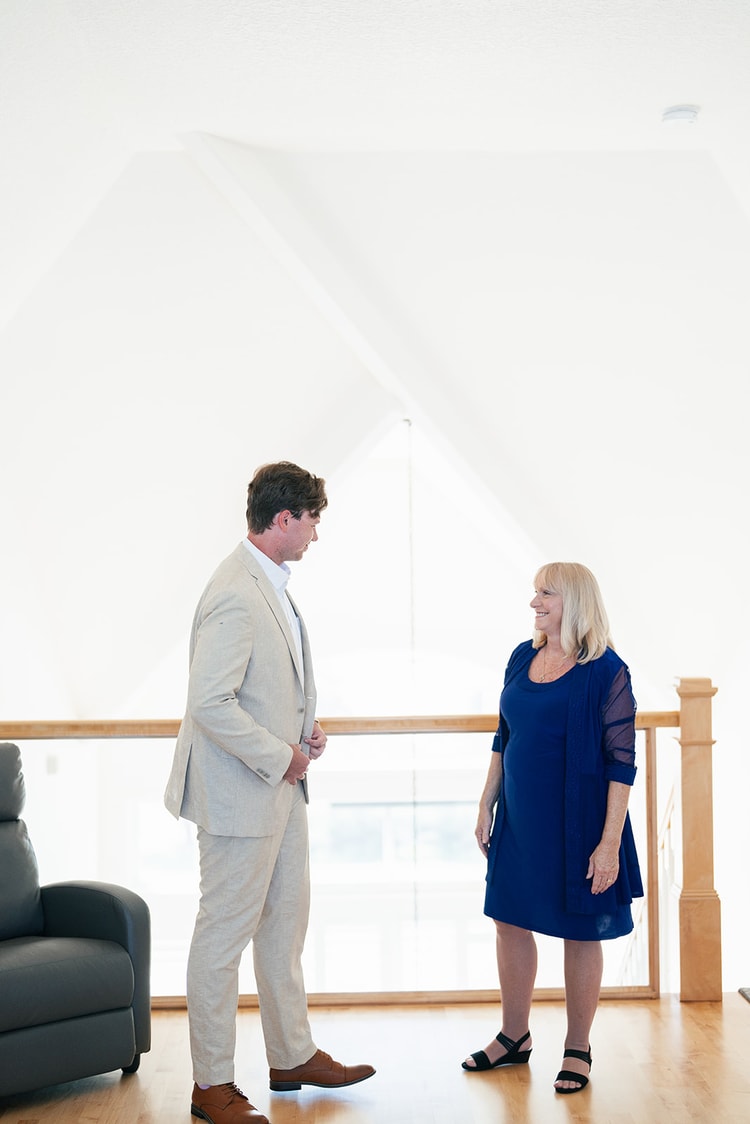 Groom in a tan suit and lavender tie chatting with a woman in a blue dress in a bright, high-ceilinged room before the ceremony.