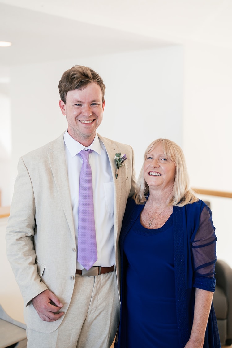 Groom in a tan suit with a lavender tie and floral boutonniere smiling alongside a woman in a royal blue dress before the ceremony.