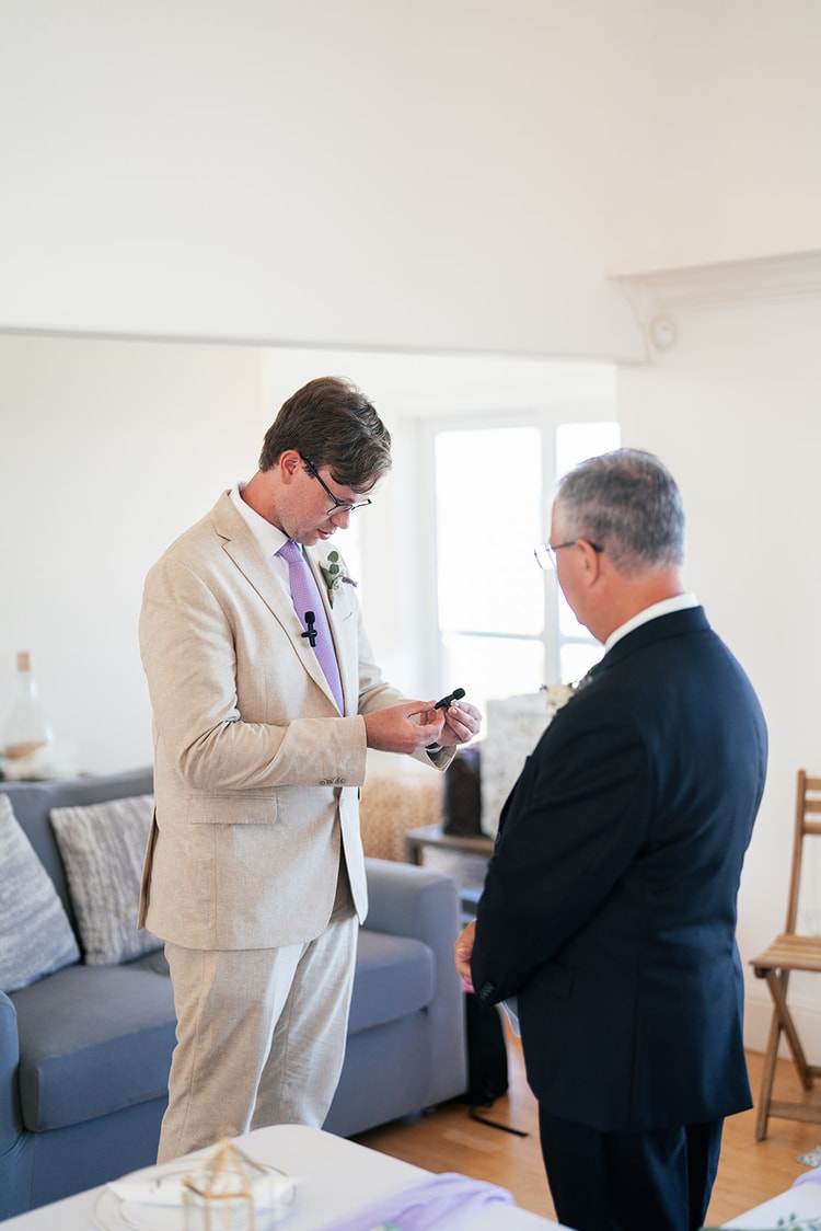 Groom in a tan suit and lavender tie looking at a lapel microphone while a man in a dark suit looks on during pre-ceremony preparations.