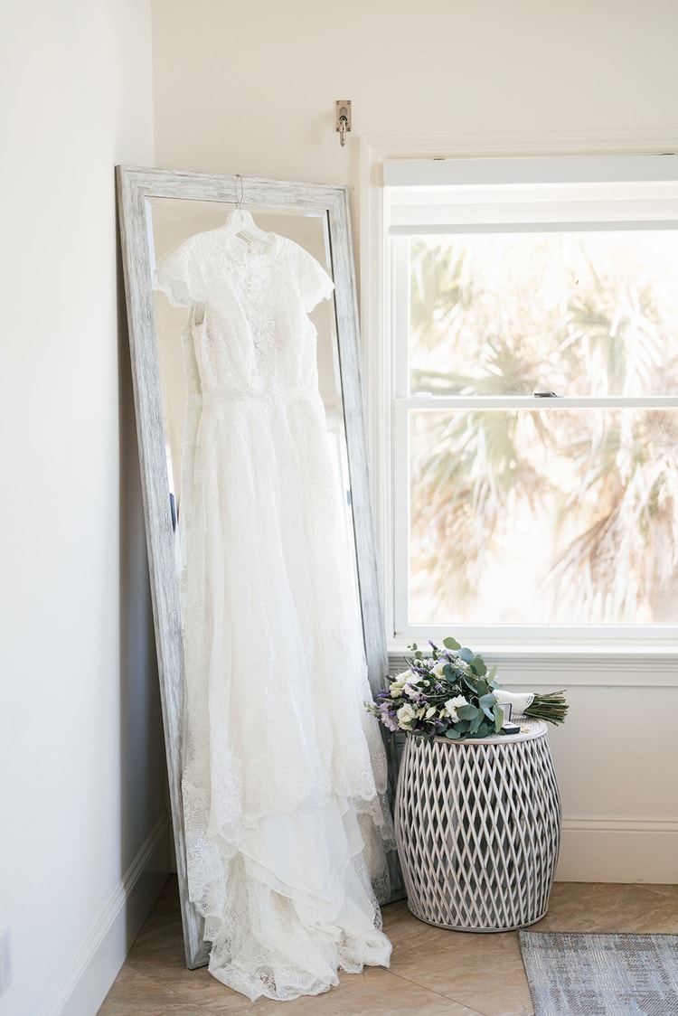 Lace wedding gown hanging from a leaning mirror beside a bright window with a palm tree visible outside; the bridal bouquet rests on a woven stool below.