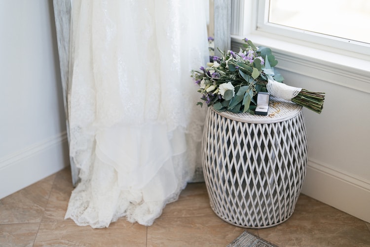 The hem and train of a lace wedding gown pooled beside a leaning mirror; the bridal bouquet and a ring box sit on a woven side table nearby.