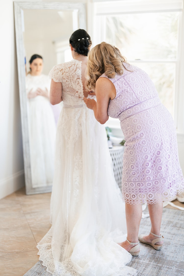 Woman in a lavender lace dress fastening the back buttons of the bride's lace wedding gown during getting-ready preparations in front of a floor mirror.