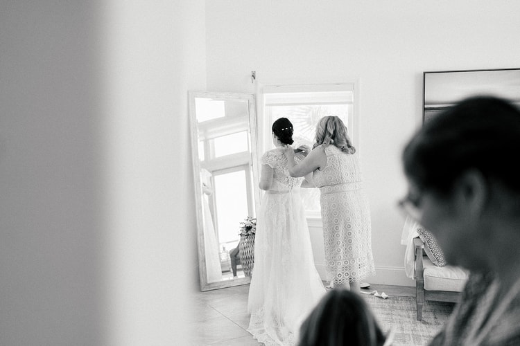 Black-and-white candid of a bride having the back of her lace gown fastened by a woman in a lace dress, watched by onlookers in an airy getting-ready room.