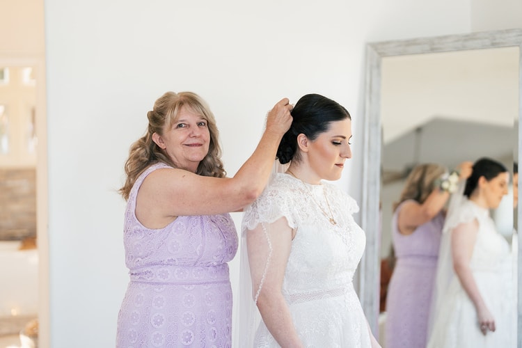 Woman in a lavender dress smiling as she pins the bride's veil into her updo, reflected in a leaning mirror during bridal prep.
