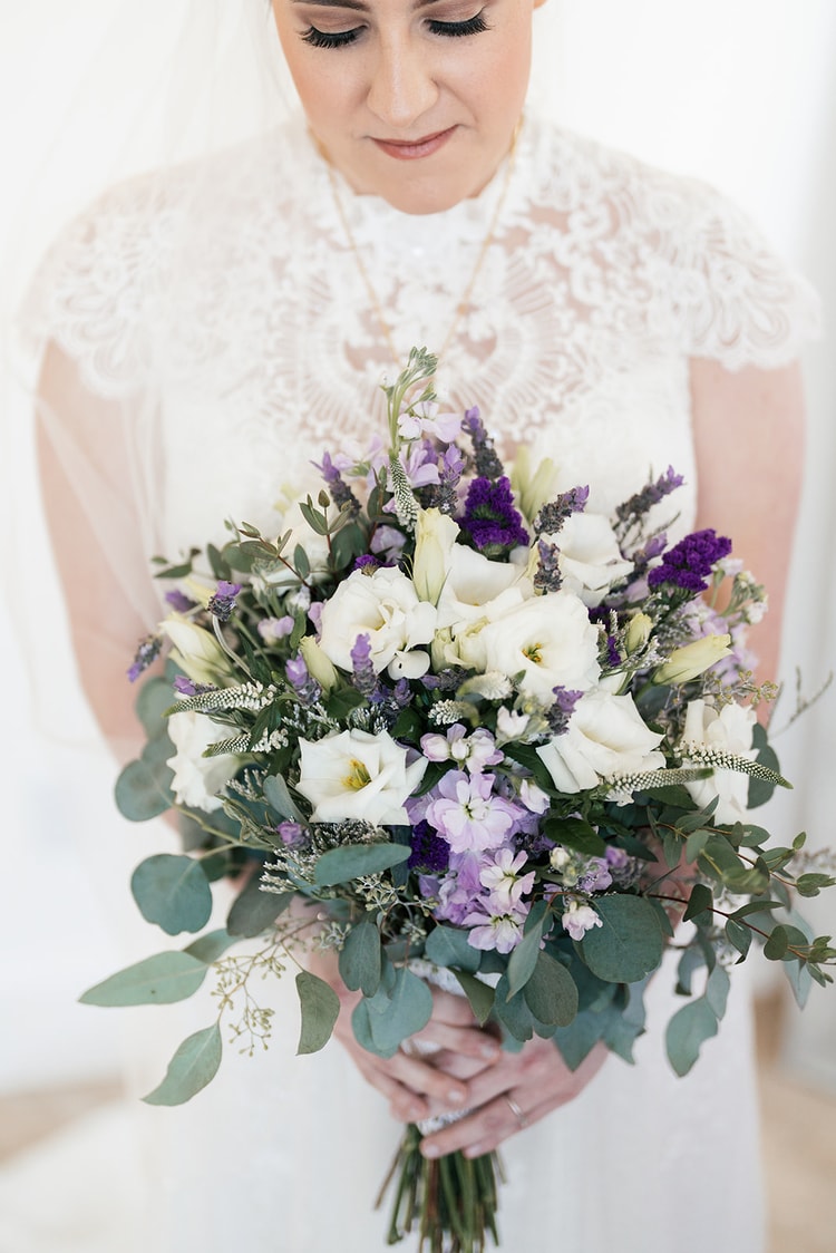 Bride looking down at her bouquet of white lisianthus, purple statice, lavender, and eucalyptus, wearing a lace cap-sleeve gown and layered gold necklace.