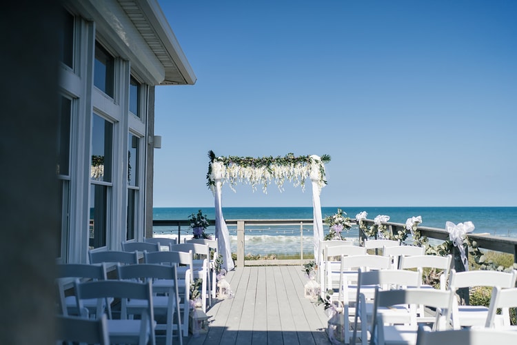Outdoor beachfront ceremony setup with white garden chairs lining a wooden deck aisle leading to a flower-draped arch, ocean and blue sky in the background.