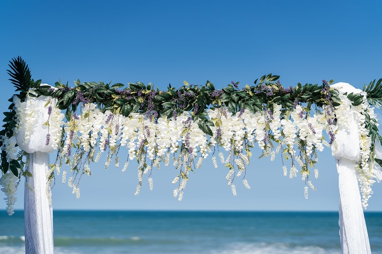 Ceremony arch draped in cascading white wisteria and green garland against a vivid blue sky and ocean backdrop.
