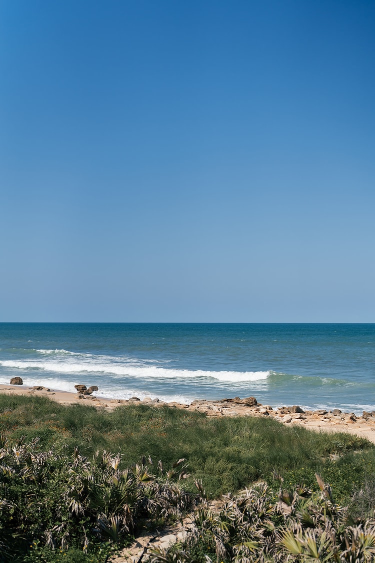 Coastal landscape of gentle surf breaking over a sandy, rock-strewn shoreline beneath a clear blue sky.