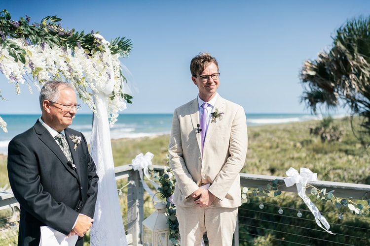 Groom standing beneath the floral ceremony arch at the ocean's edge, smiling warmly as he watches the bride approach.