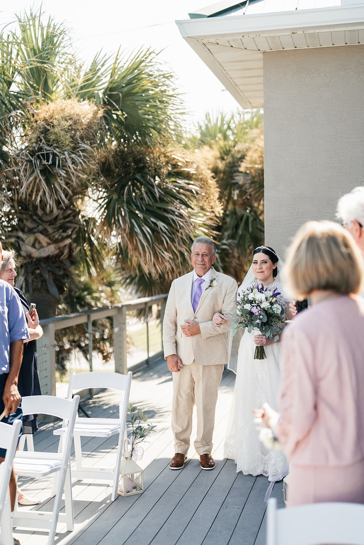 Bride walking down the aisle on the arm of her escort, holding a lush purple and white bouquet, with palm trees and seated guests behind her.