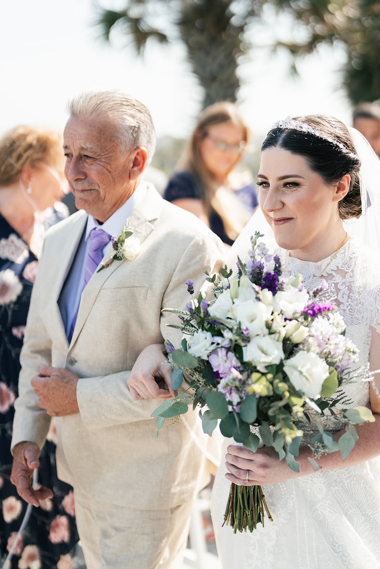 Close portrait of the bride walking up the aisle with her escort, gazing ahead calmly as she holds her purple and white bouquet.