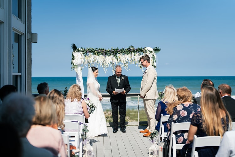 Wide shot of the outdoor beach ceremony: bride and groom face each other under the floral arch while the officiant reads aloud and guests look on.