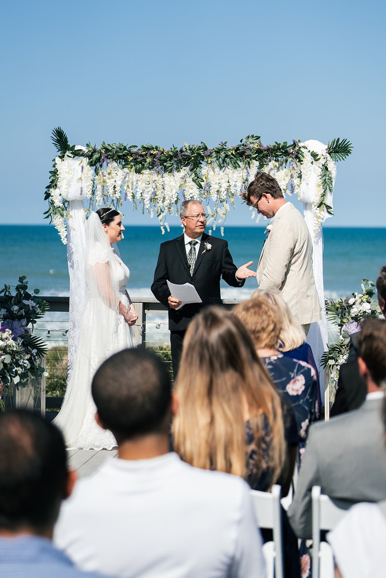 Groom reading his vows from a card under the ceremony arch as the bride watches and guests observe from the seated rows behind.