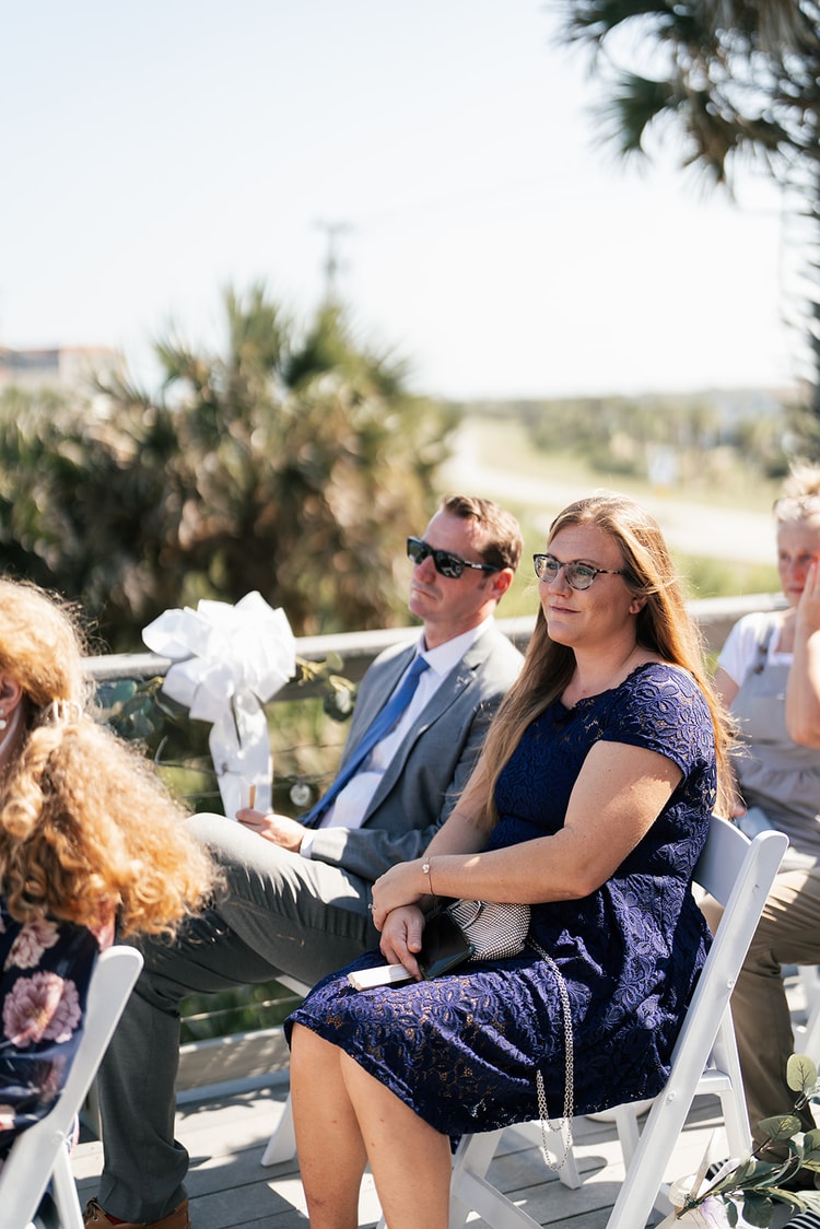 Wedding guests seated on white folding chairs at the outdoor beach ceremony, watching the proceedings with attention.