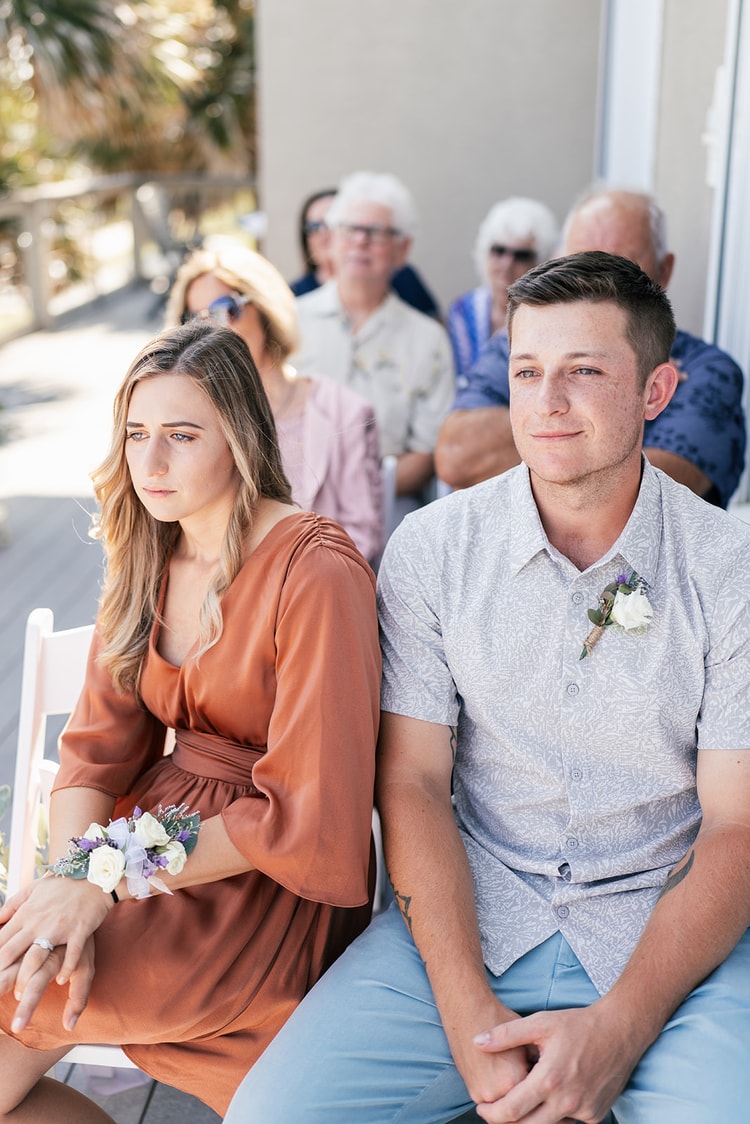 Two young guests seated in the front row at the outdoor ceremony; the young man wears a boutonniere and the young woman a floral wrist corsage.